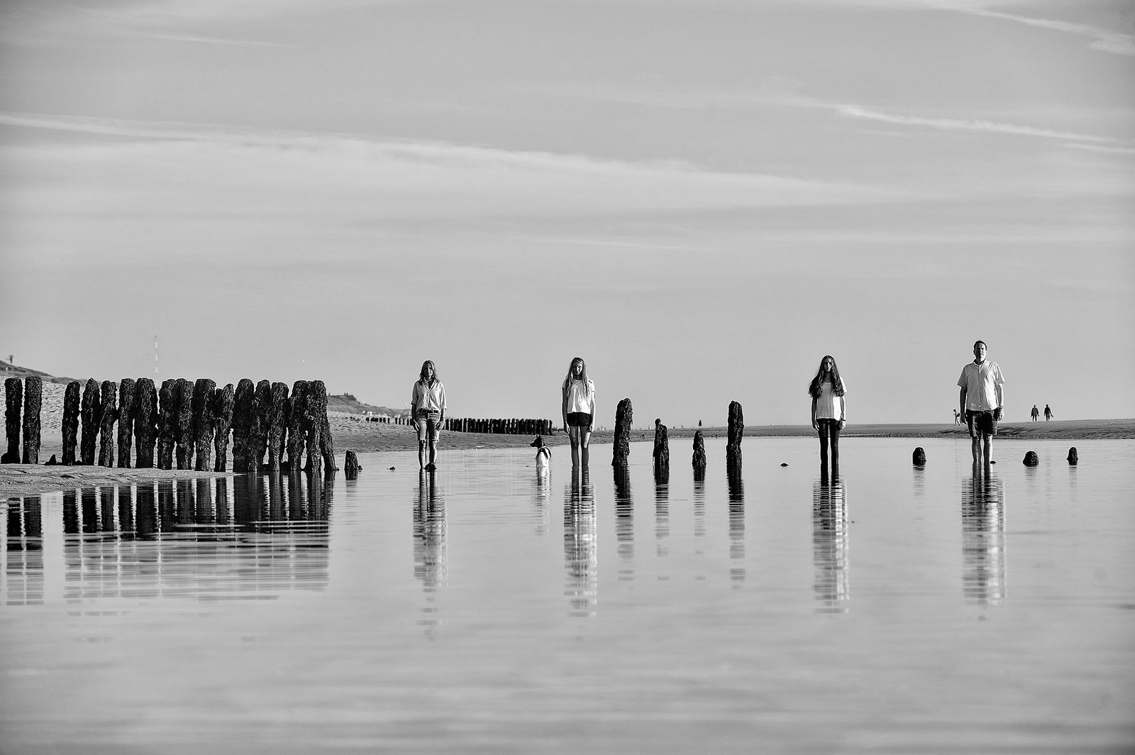 Eine vierköpfige Familie steht starr und gerade im flachen Wasser der Sandbank auf der Westküste von Sylt, ihres Spiegelbild sieht man in der Meeroberfläche. Die Buhnen kompletieren das Bild